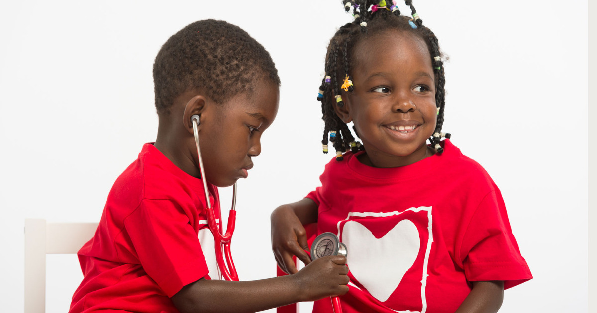Two smiling children wearing heart logo shirts playing with a stethoscope, symbolizing the importance of children’s heart surgery in Kenya.