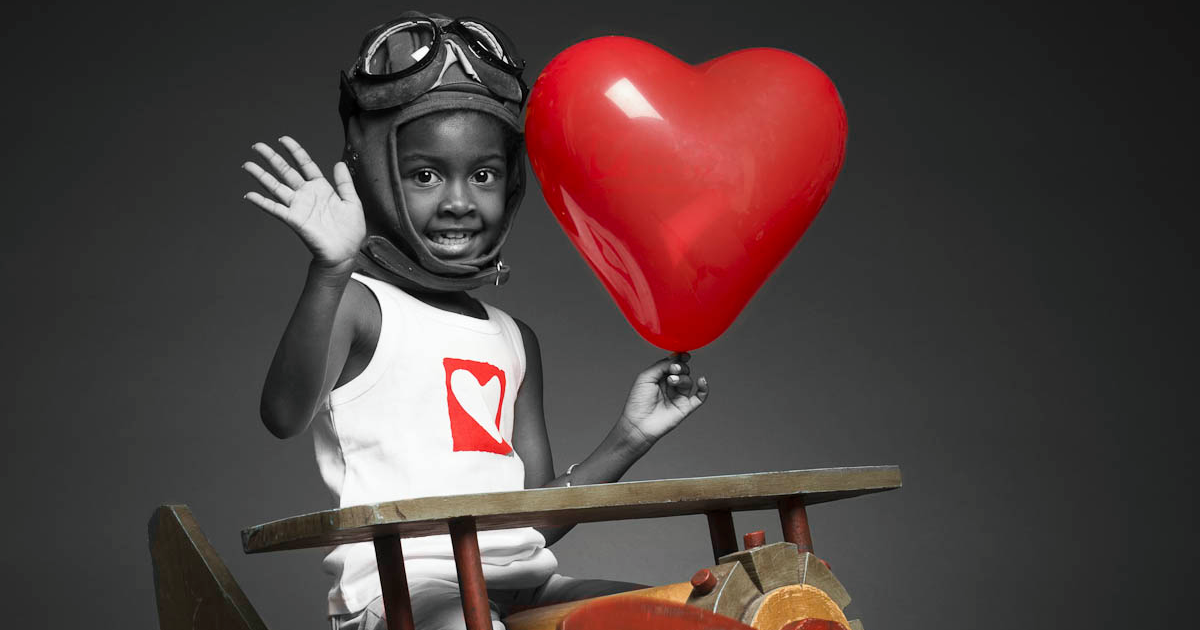 Smiling child in a heart-logo shirt holding a red heart balloon, symbolizing hope after children’s heart surgery in Kenya.